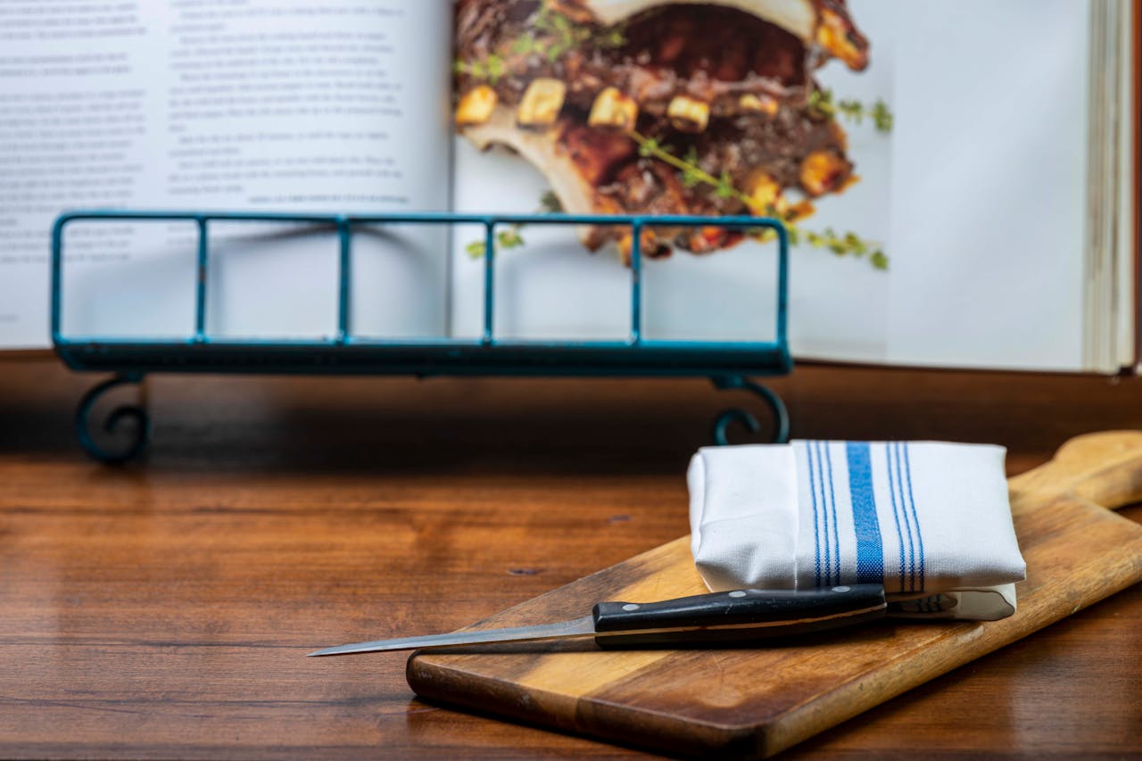 Close-up of a knife on a cutting board, with a cookbook in a kitchen setting.